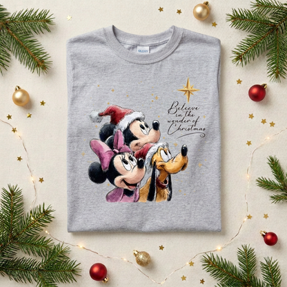 A folded sport gray T-shirt with a Mickey Mouse, Minnie Mouse, and Pluto Christmas graphic, styled in a flat lay with pine branches, gold ornaments, and string lights on a light beige background.