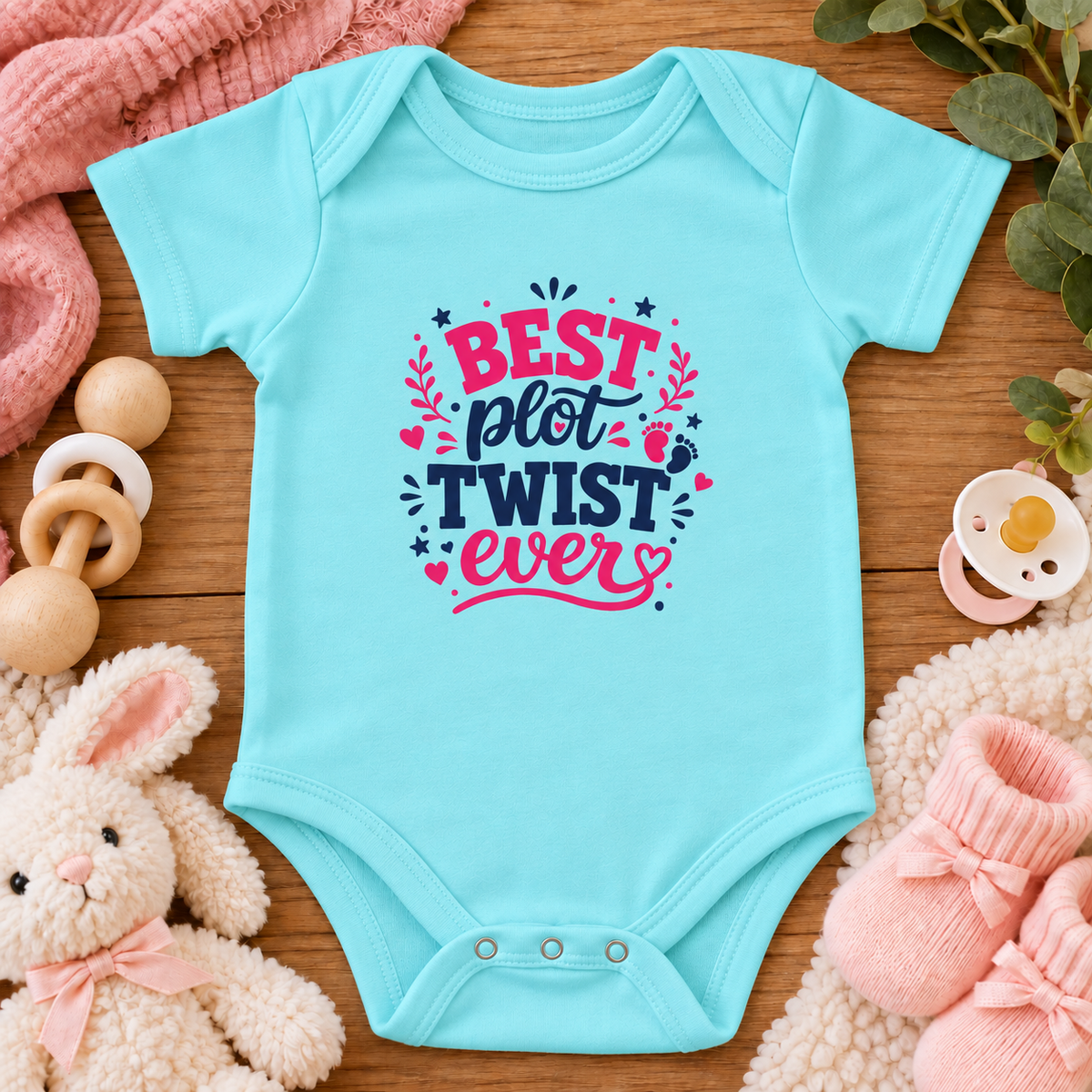 A chill blue baby bodysuit with a colorful graphic reading 'BEST plot TWIST ever' in pink and navy lettering, styled in a flat lay on a wooden surface with a pink blanket, wooden toy, pacifier, bunny plush, and pink booties.