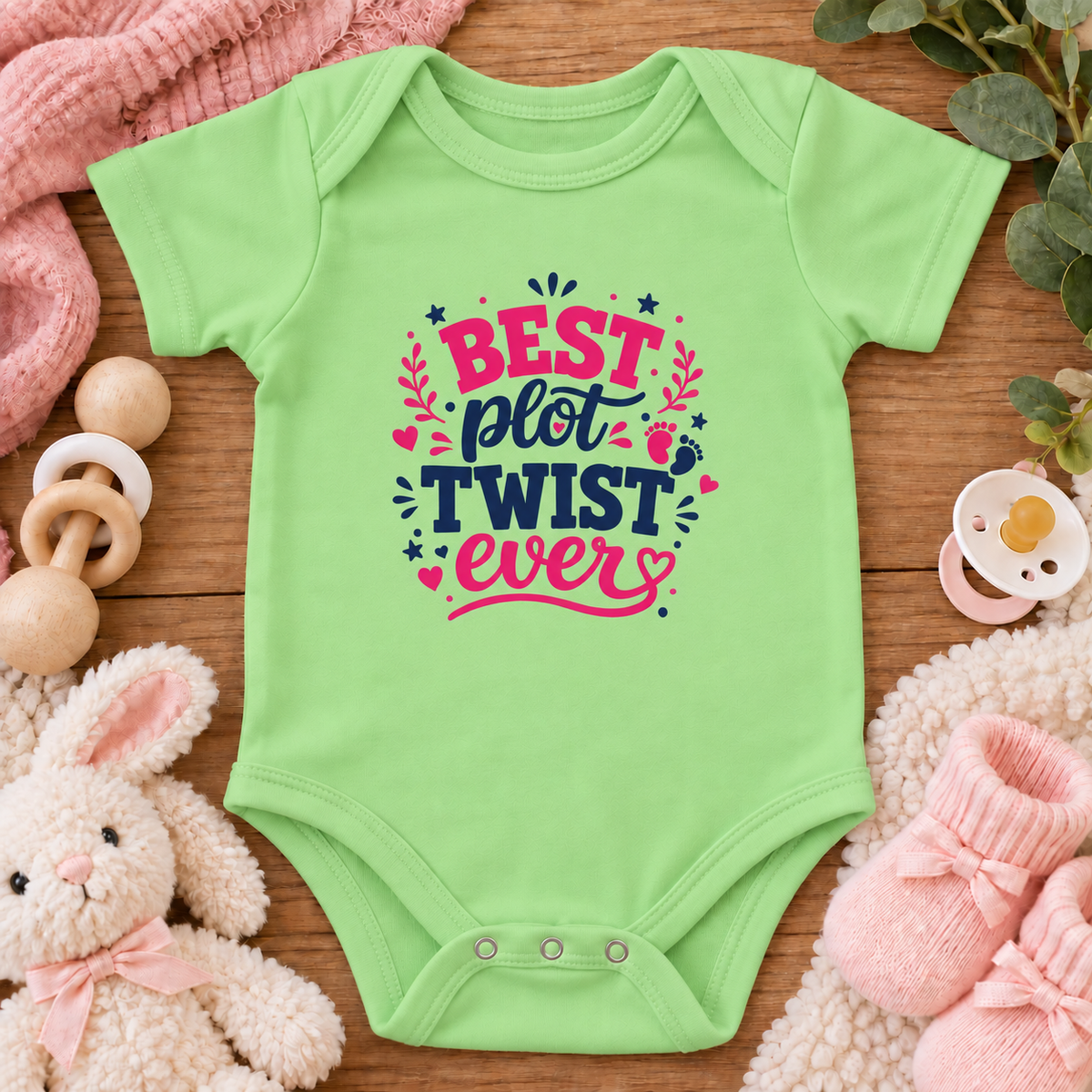 A keylime green baby bodysuit with a colorful graphic that reads 'BEST plot TWIST ever' in pink and navy lettering, surrounded by baby-themed props including a stuffed bunny, pink socks, a wooden pacifier clip, and a pink blanket on a wooden surface.