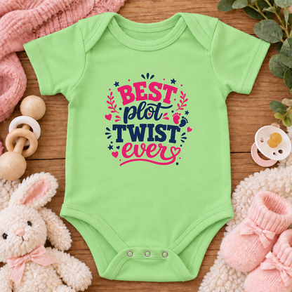 A keylime green baby bodysuit with a colorful graphic that reads 'BEST plot TWIST ever' in pink and navy lettering, surrounded by baby-themed props including a stuffed bunny, pink socks, a wooden pacifier clip, and a pink blanket on a wooden surface.