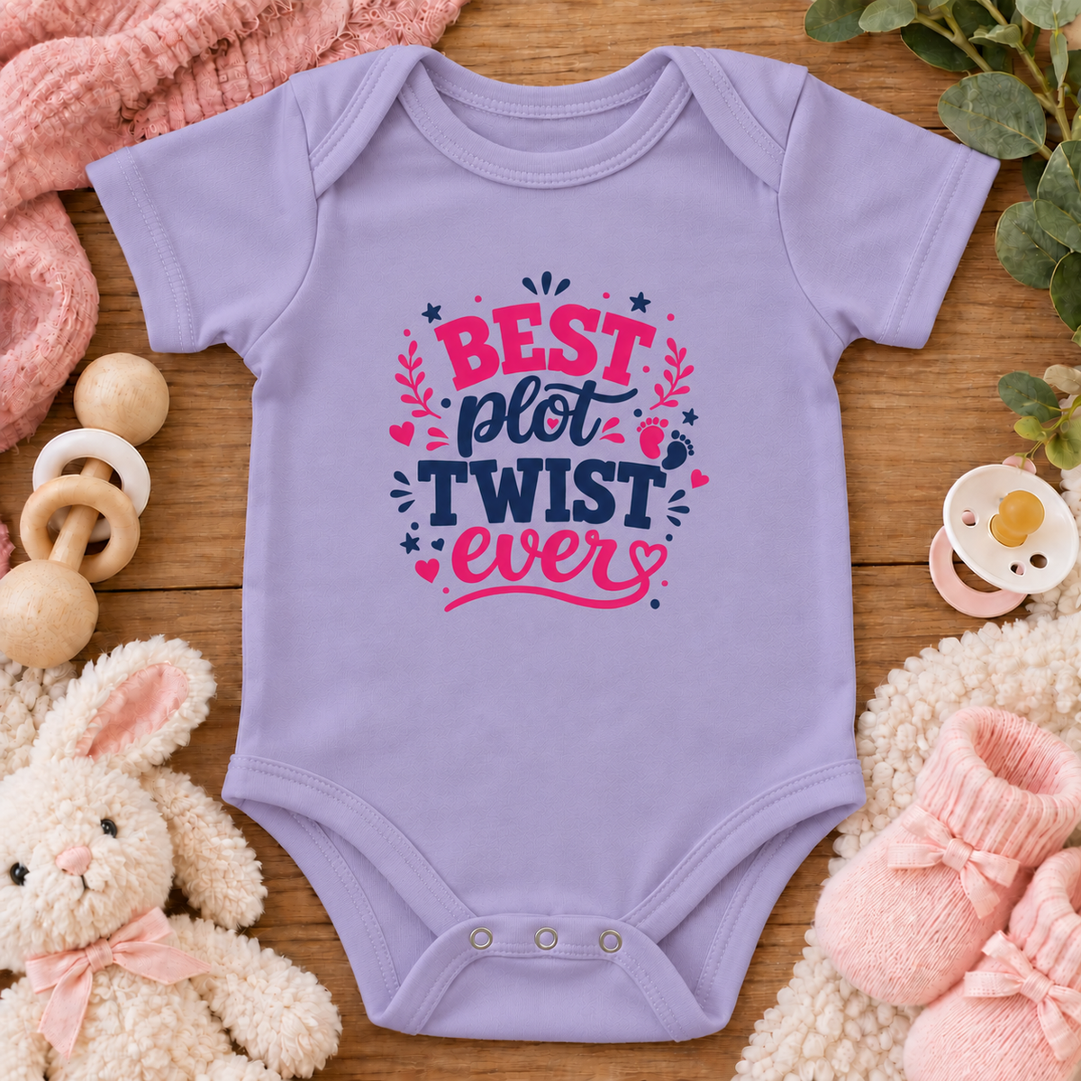 A lavender baby bodysuit with a colorful graphic reading 'BEST plot TWIST ever' in pink and navy, styled in a flat lay on a wooden surface with a plush bunny, pink knit socks, a pacifier, a wooden teething ring, and a soft blanket.