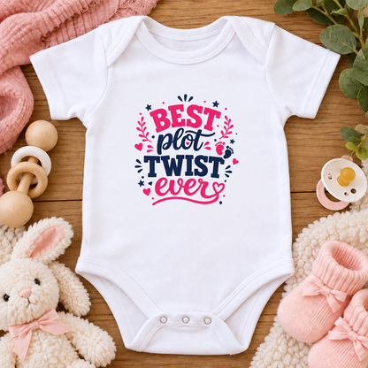 A white baby bodysuit laid flat on a wooden surface, surrounded by baby-themed props including a plush bunny, pink knit socks, a pacifier, wooden teethers, and a soft blanket. The bodysuit has a colorful printed slogan reading “BEST plot TWIST ever” in pink and navy lettering with small decorative accents.