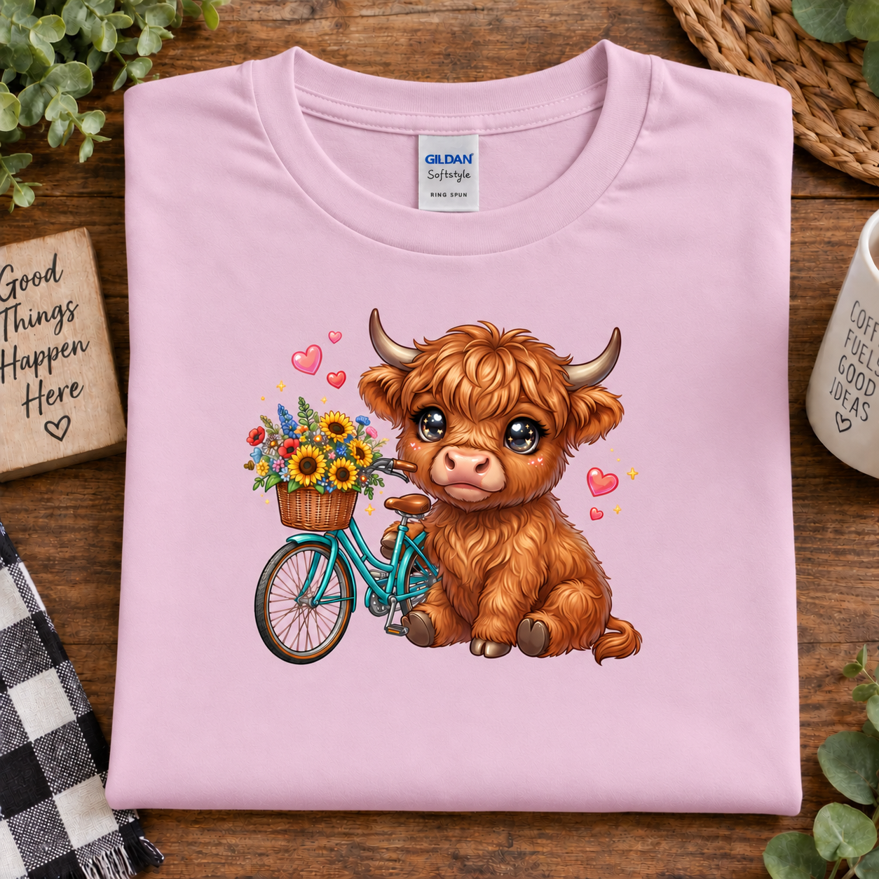 A folded light pink Gildan Softstyle T-shirt is displayed on a wooden surface with decorative props. The shirt features a colorful cartoon graphic of a fluffy brown Highland cow sitting on a teal bicycle with a basket of sunflowers, surrounded by small pink hearts.