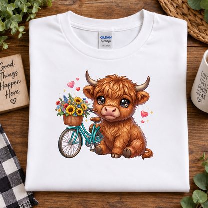 A folded white Gildan Softstyle T-shirt with a cute cartoon illustration of a fluffy brown Highland cow riding a teal bicycle with a basket of sunflowers, placed on a wooden surface with decorative props.