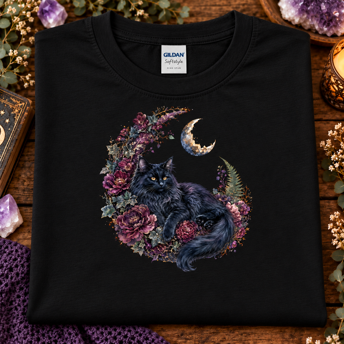 A folded black Gildan Softstyle t-shirt features a graphic of a fluffy black cat curled up inside a crescent moon wreath decorated with purple and pink flowers.