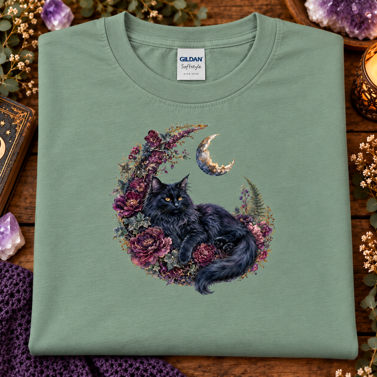 A folded sage green Gildan Softstyle t-shirt features a graphic of a fluffy black cat curled up inside a crescent moon wreath decorated with purple and pink flowers.