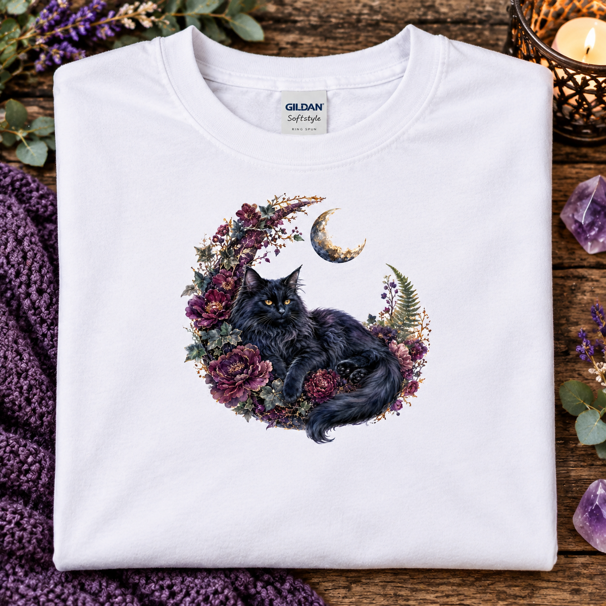 A folded white Gildan Softstyle t-shirt features a graphic of a fluffy black cat curled up inside a crescent moon wreath decorated with purple and pink flowers.