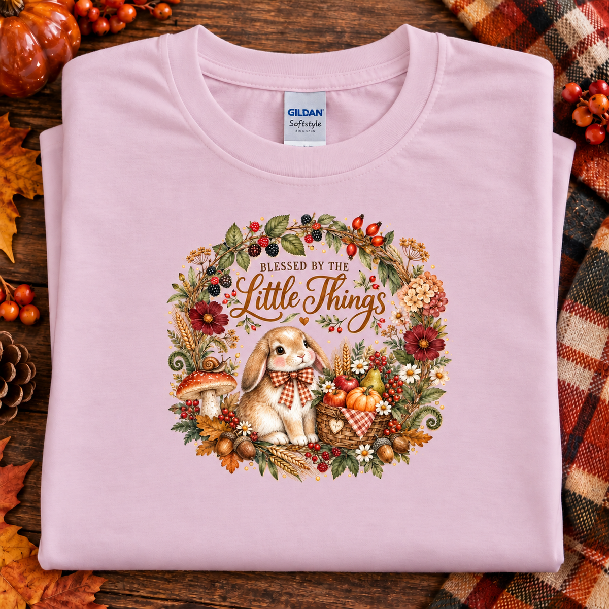 A folded light pink Gildan Softrusty t-shirt with a detailed autumn-themed graphic of a bunny in a floral wreath and the text 'BLESSED BY THE Little Things'. The shirt is styled on a rustic wooden background with fall leaves, pumpkins, and a plaid blanket.