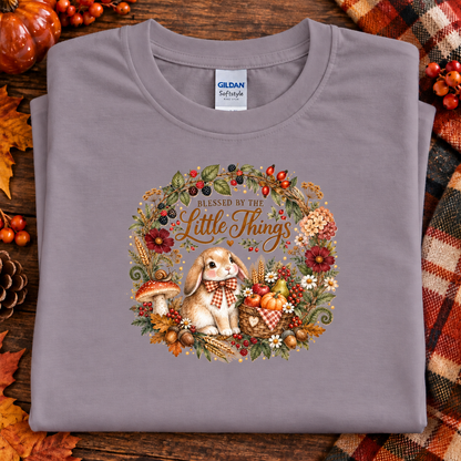 A folded paragon Gildan Softstyle T-shirt with a detailed autumn-themed graphic of a bunny in a floral wreath and the text 'Blessed by the Little Things,' styled on a wooden surface with fall leaves, pumpkins, and a plaid blanket.