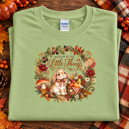 A folded pistachio green Gildan Softstyle T-shirt with a detailed autumn-themed bunny graphic and the text 'BLESSED BY THE Little Things,' displayed on a rustic wooden background with fall leaves, pumpkins, and a plaid blanket.