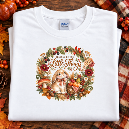 A folded white Gildan Softstyle T-shirt with a large autumn-themed bunny graphic and the phrase 'Blessed by the Little Things,' displayed on a wooden surface with fall decor and a plaid blanket.