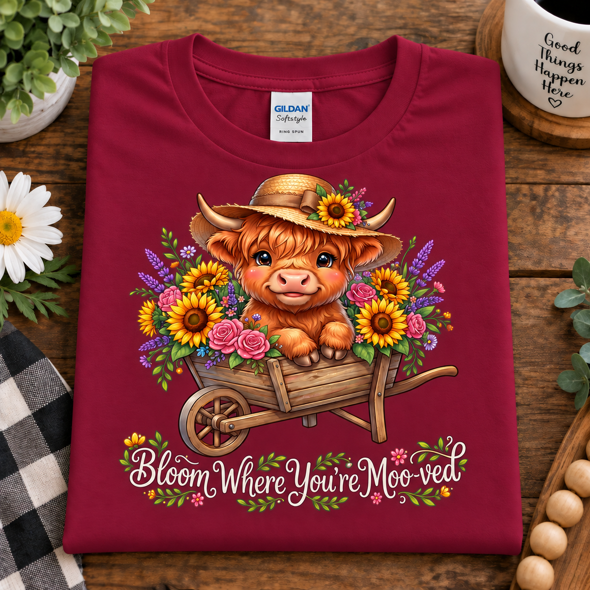 A folded cardinal red t-shirt with a colorful graphic of a Scottish Highland cow in a wheelbarrow surrounded by flowers, with the text 'Bloom Where You're Moo'd' underneath. The shirt is displayed on a wooden table with decorative props including a daisy, a mug, and a plant.