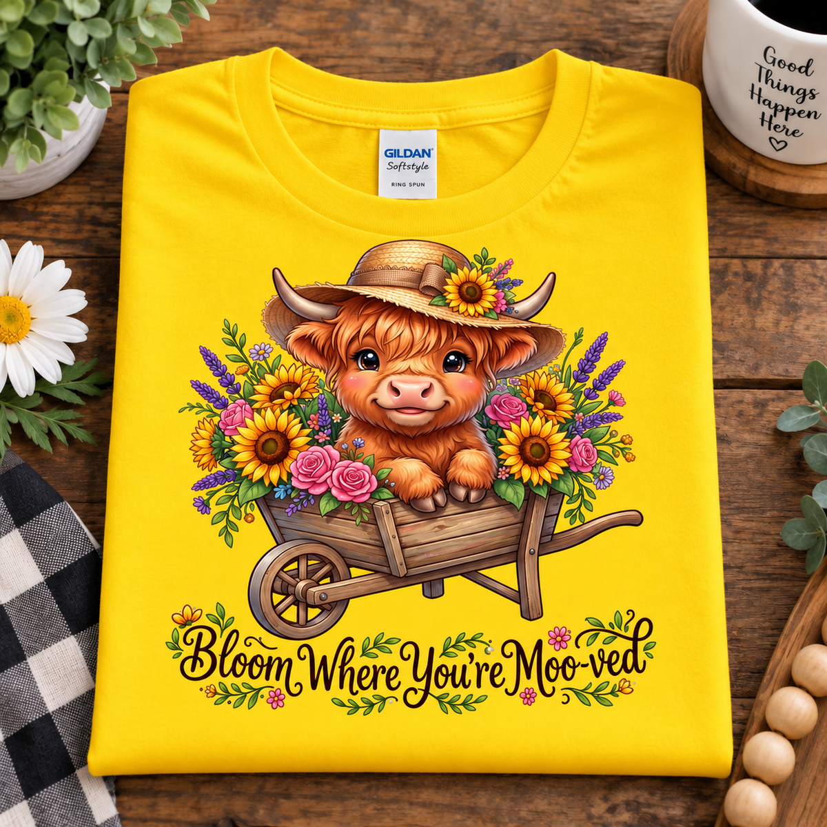 A folded daisy colored Gildan Softstyle T-shirt is displayed on a rustic wooden surface. The shirt features a colorful graphic of a highland cow in a straw hat sitting in a wheelbarrow filled with sunflowers and roses, with the text 'Bloom Where You're MooVED' printed below.