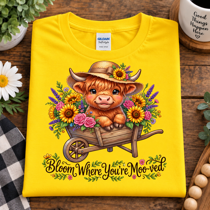 A folded daisy colored Gildan Softstyle T-shirt is displayed on a rustic wooden surface. The shirt features a colorful graphic of a highland cow in a straw hat sitting in a wheelbarrow filled with sunflowers and roses, with the text 'Bloom Where You're MooVED' printed below.