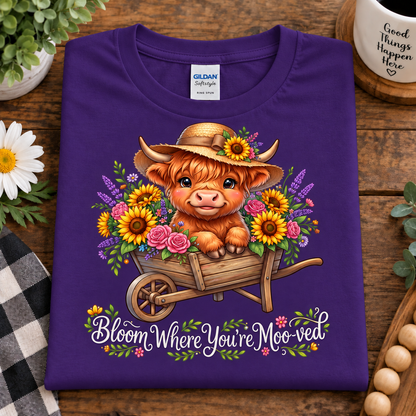 A folded purple Gildan Softstyle T-shirt with a colorful graphic of a fluffy orange cow in a straw hat and flower cart, with the text 'Bloom Where You're Moo'd' on a wooden surface.