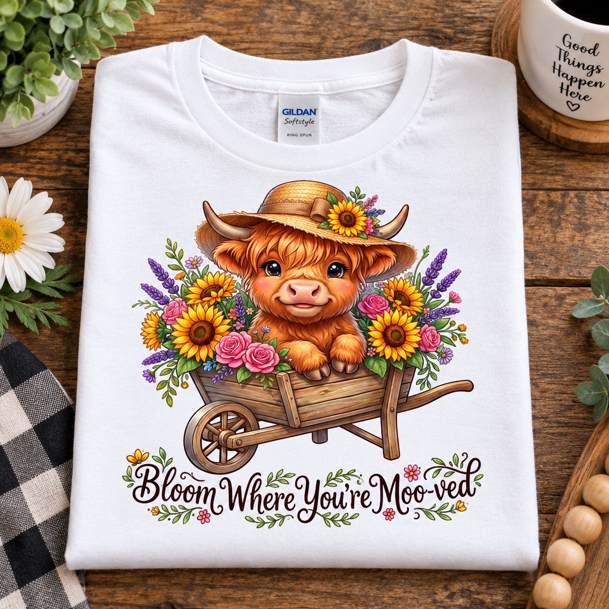 A folded white Gildan Softstyle T-shirt is displayed on a rustic wooden surface. The shirt features a colorful graphic of a fluffy brown Scottish highland cow sitting in a wooden wheelbarrow filled with yellow sunflowers, pink roses, and purple lavender.