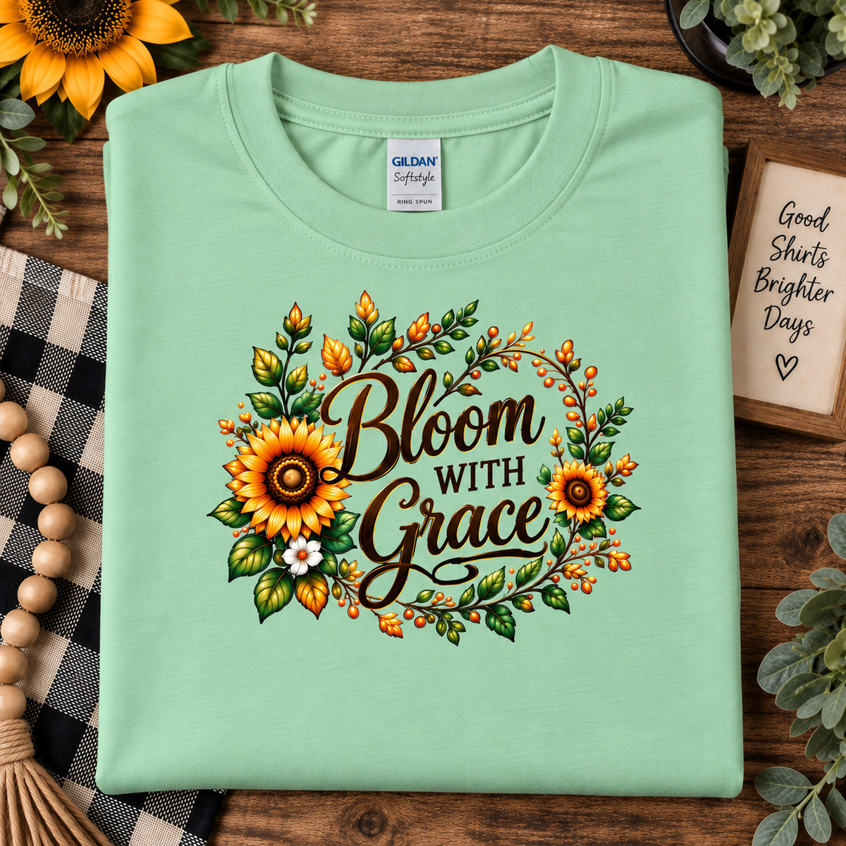 A folded mint green Gildan Softstyle t-shirt is displayed on a rustic wooden background with autumn-themed props like sunflowers, a plaid blanket, and a wooden sign. The shirt features a 'Bloom With Grace' graphic surrounded by sunflowers, green leaves, and orange berries.