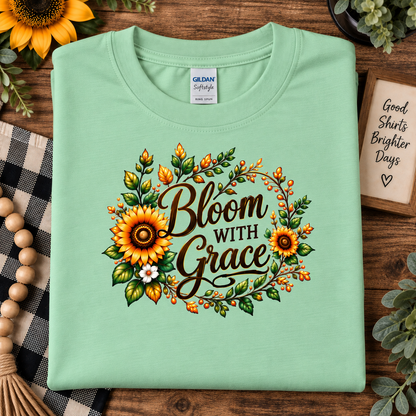 A folded mint green Gildan Softstyle t-shirt is displayed on a rustic wooden background with autumn-themed props like sunflowers, a plaid blanket, and a wooden sign. The shirt features a 'Bloom With Grace' graphic surrounded by sunflowers, green leaves, and orange berries.