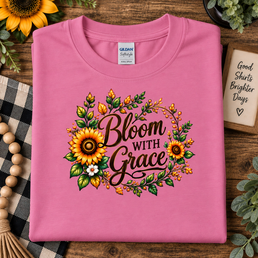 A folded azalea pink Gildan Softstyle t-shirt with a 'Bloom With Grace' floral graphic, displayed on a rustic wooden background with sunflowers, a checkered cloth, and a small sign.