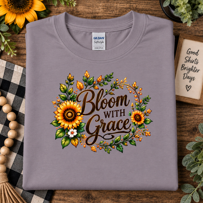 A folded light paragon purple-gray Gildan Softstyle t-shirt with a 'Bloom With Grace' sunflower graphic, styled on a rustic wooden background with decorative props.