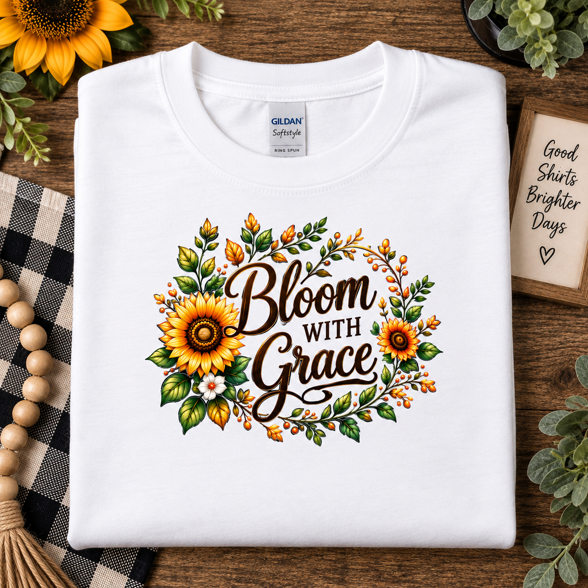 A folded white Gildan Softstyle T-shirt with a 'Bloom With Grace' sunflower graphic, styled on a wooden surface with decorative props.