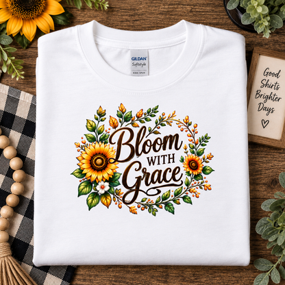 A folded white Gildan Softstyle T-shirt with a 'Bloom With Grace' sunflower graphic, styled on a wooden surface with decorative props.