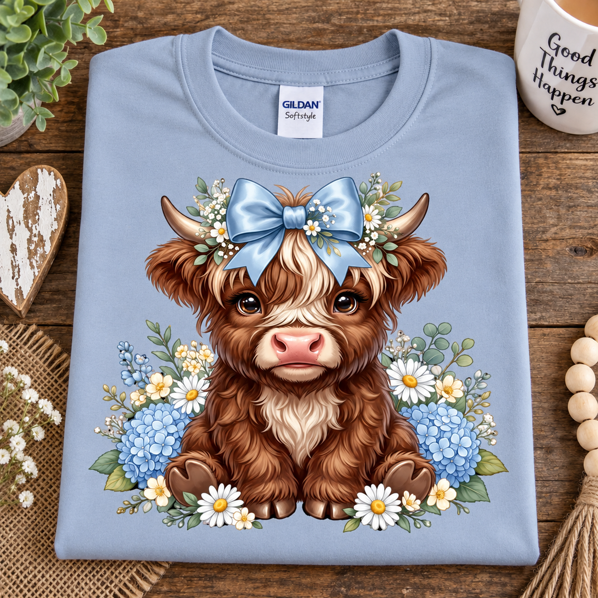 A folded light blue Gildan Softstyle t-shirt with a detailed graphic of a cute brown and white highland cow wearing a blue bow and floral crown, displayed on a rustic wooden background with decorative props.