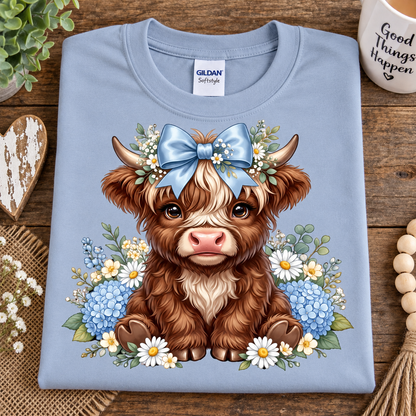 A folded light blue Gildan Softstyle t-shirt with a detailed graphic of a cute brown and white highland cow wearing a blue bow and floral crown, displayed on a rustic wooden background with decorative props.