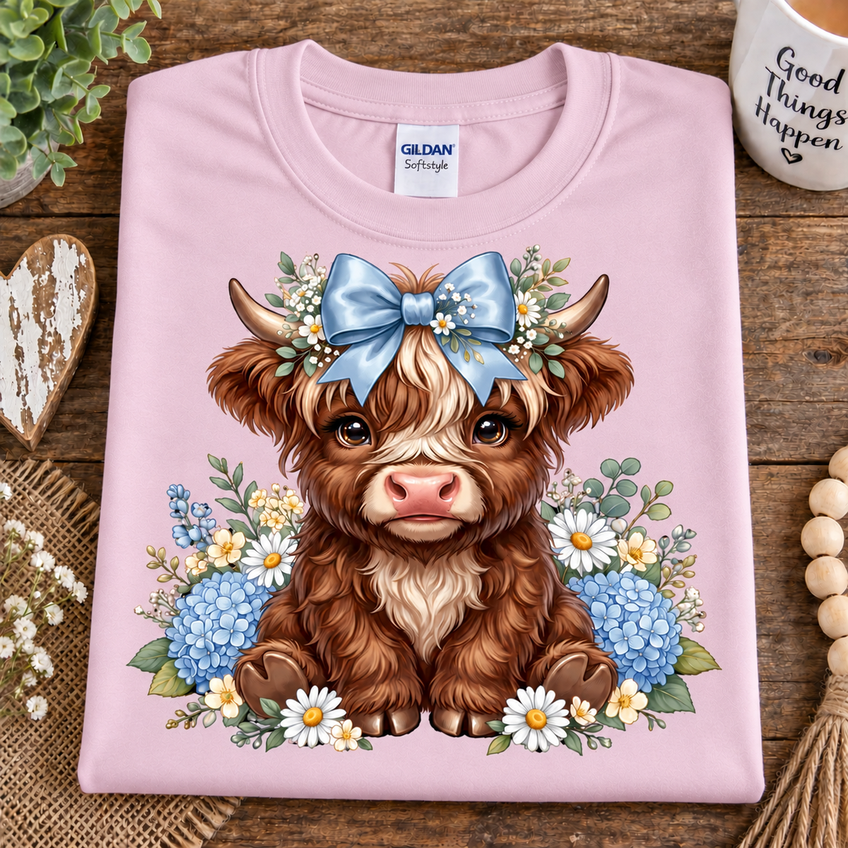 A folded light pink Gildan Softstyle t-shirt with a detailed graphic of a cute brown Scottish highland cow wearing a blue bow and floral crown, displayed on a rustic wooden background with decorative props.