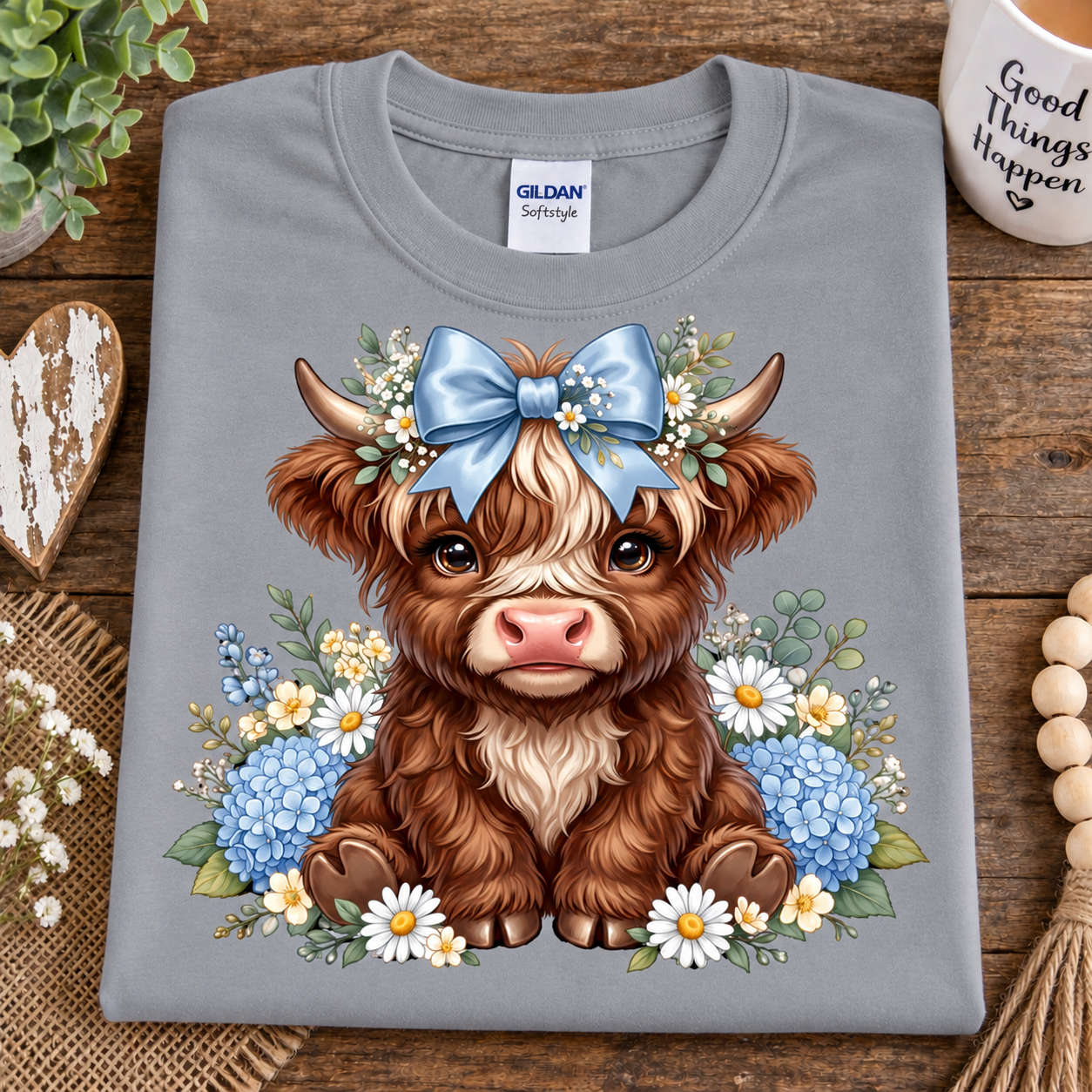A folded sport gray Gildan Softee t-shirt with a cute cartoon Highland cow graphic, styled on a rustic wooden background with decorative props.