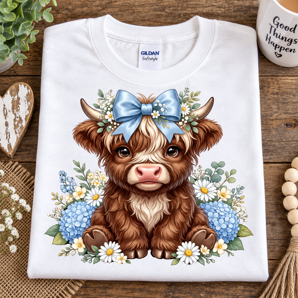 A folded white Gildan Softstyle T-shirt with a detailed graphic of a brown highland cow wearing a blue bow and floral crown, displayed on a rustic wooden surface with decorative props.