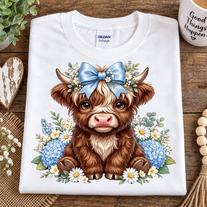 A folded white Gildan Softstyle T-shirt with a detailed graphic of a brown highland cow wearing a blue bow and floral crown, displayed on a rustic wooden surface with decorative props.
