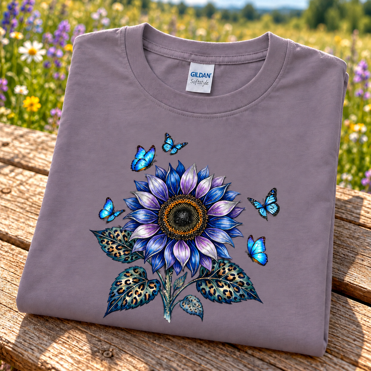 A folded Gildan Softstyle t-shirt in paragon, featuring a vibrant graphic of a blue and purple sunflower with blue butterflies and leopard-print leaves, displayed on a rustic wooden bench with a blurred meadow background.