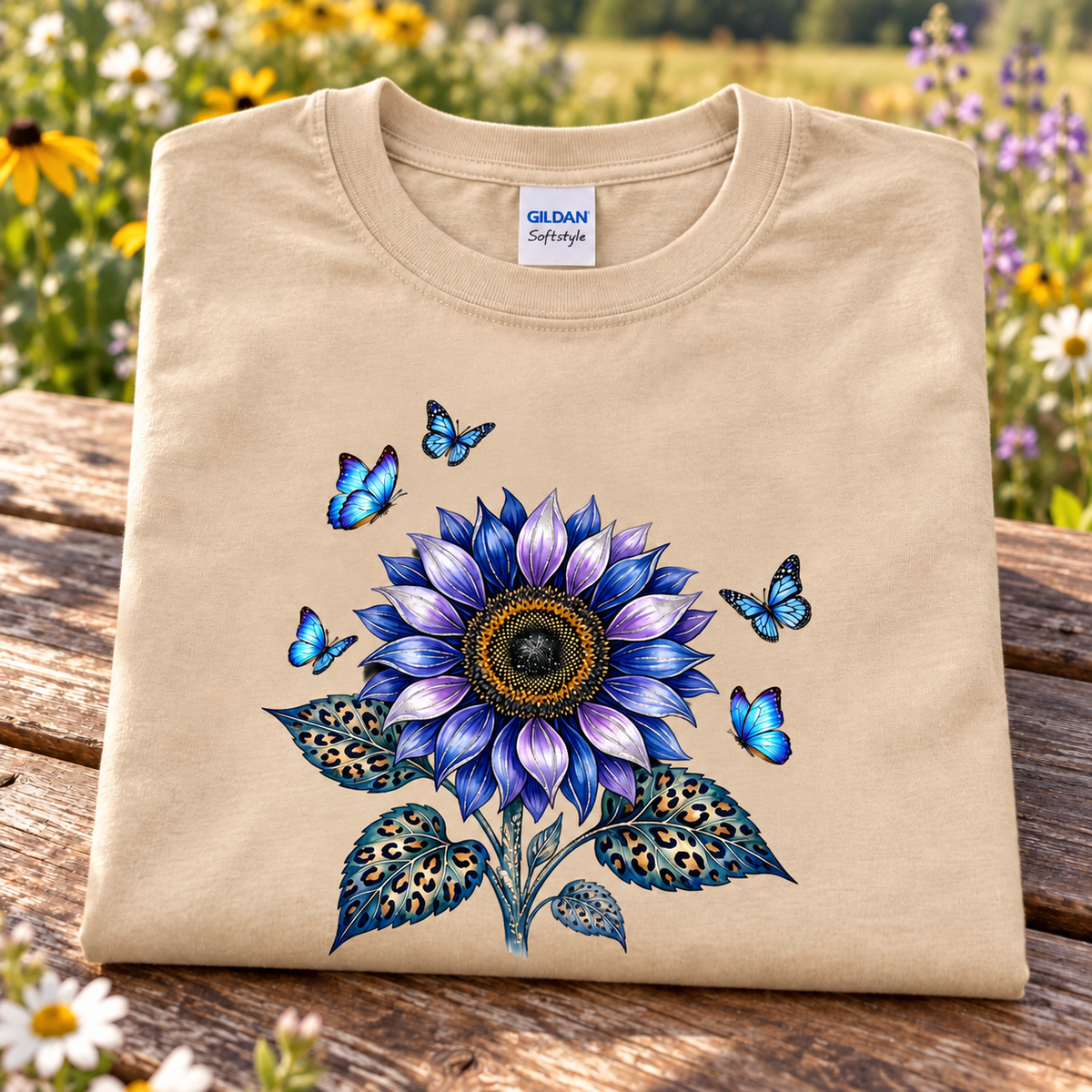 A sand Gildan Softstyle t-shirt is folded and displayed on a rustic wooden table with a blurred background of a flower field. The shirt features a large graphic print of a blue and purple sunflower with matching blue butterflies.