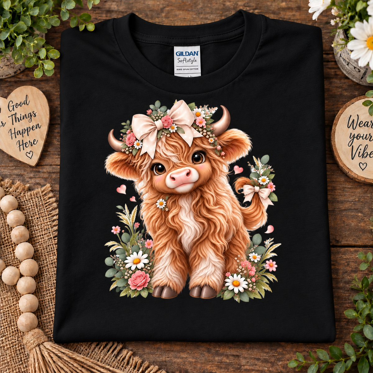 A folded black Gildan Softstyle t-shirt with a detailed graphic of a fluffy brown highland cow surrounded by a floral crown and flowers, displayed on a rustic wooden background with decorative props.