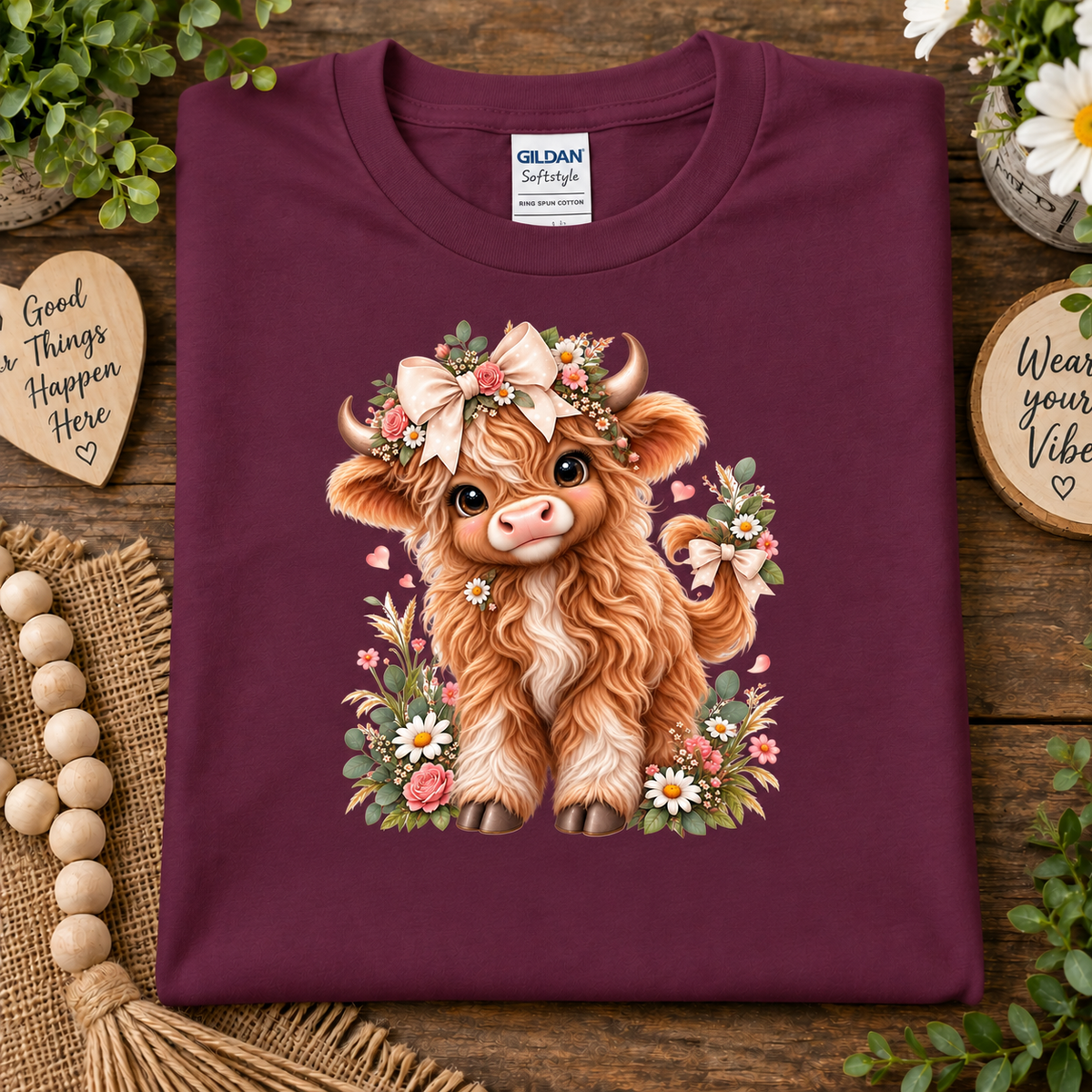 A folded maroon Gildan Softstyle t-shirt is displayed on a rustic wooden surface. The shirt features a detailed graphic of a fluffy brown highland cow decorated with a floral crown and surrounded by pink and white flowers.