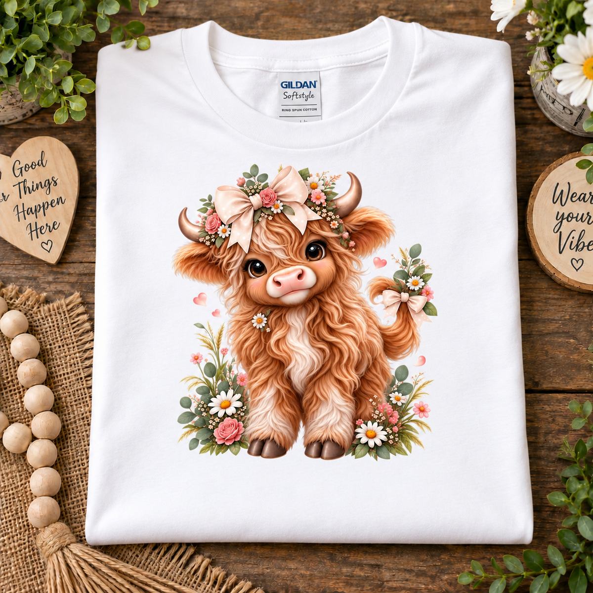 A folded white Gildan Softstyle T-shirt with a colorful graphic of a fluffy brown cow with a floral crown and flowers, displayed on a rustic wooden surface with decorative props.