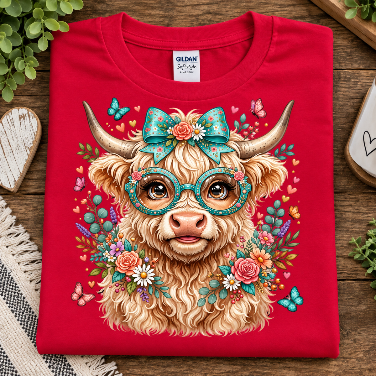 A folded red Gildan Softstyle T-shirt with a colorful graphic of a fluffy light-brown horned animal wearing teal glasses and a teal bow, surrounded by flowers, butterflies, and hearts, displayed on a wooden surface with decorative props.