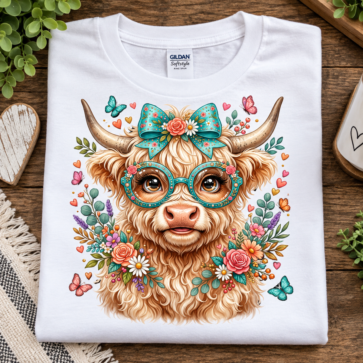 A folded white Gildan Softstyle T-shirt laid flat on a rustic wooden surface, featuring a colorful graphic of a fluffy tan animal face with large glasses, a teal bow, flowers, butterflies, and hearts.