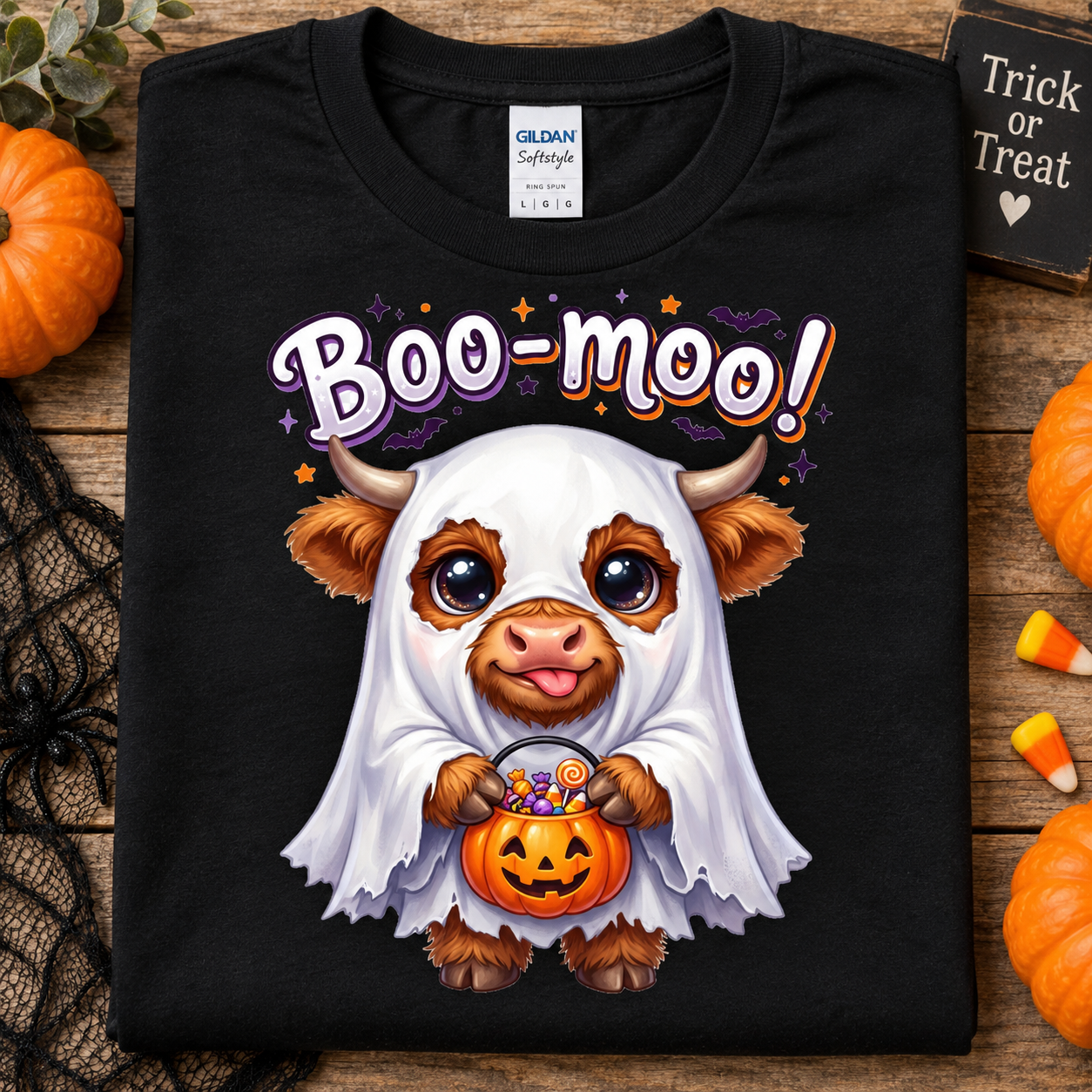A black Gildan Softstyle T-shirt laid flat on a rustic wood surface, featuring a Halloween graphic of a cute cartoon cow dressed as a ghost holding a jack-o'-lantern with candy. The shirt is surrounded by seasonal props including pumpkins, black lace, candy corn, and a small sign reading 'Trick or Treat'.