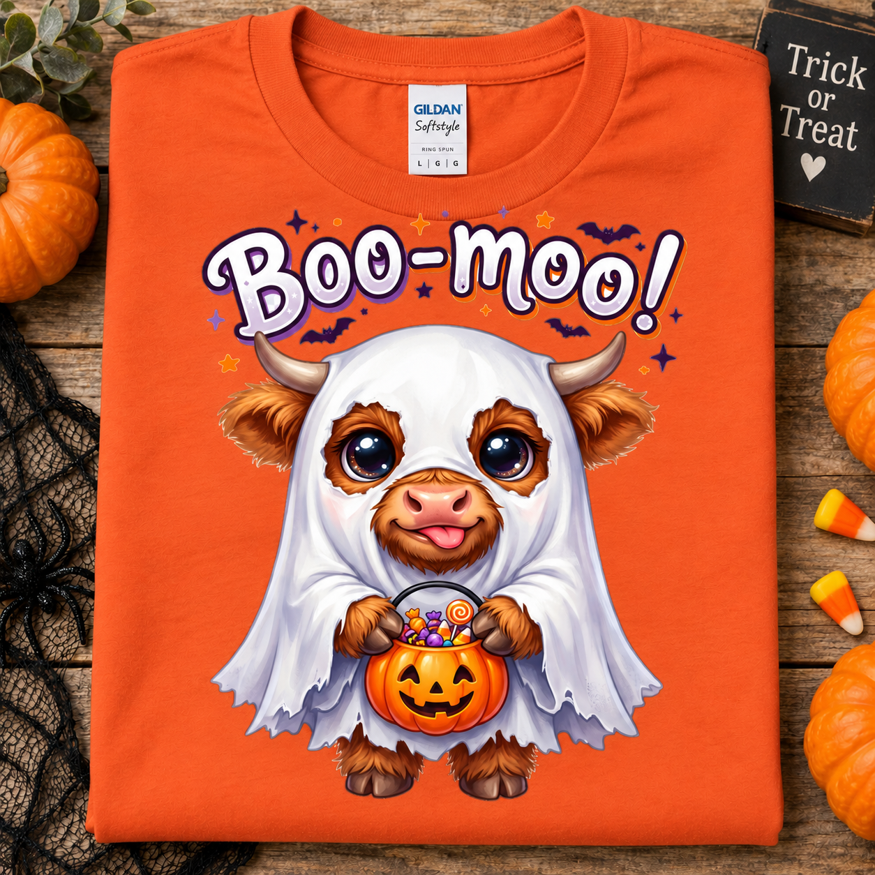 An orange Gildan Softstyle T-shirt laid flat on a rustic wood surface, featuring a cartoon cow dressed as a ghost holding a pumpkin bucket with candy, with the text 'Boo-moo!' above. Small pumpkins, candy corn, black lace, and a small sign are arranged around the shirt.
