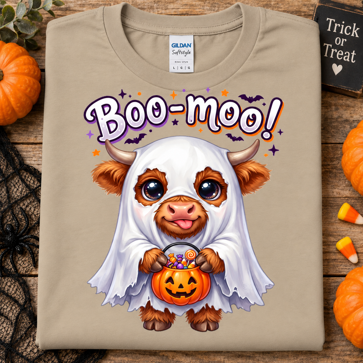 A sand Gildan Softstyle t-shirt featuring a cute cartoon cow dressed in a white ghost costume holding a jack-o'-lantern bucket of candy, with 'Boo-moo!' written above. The shirt is displayed flat on a rustic wooden background with Halloween props like pumpkins, black lace, and candy corn.