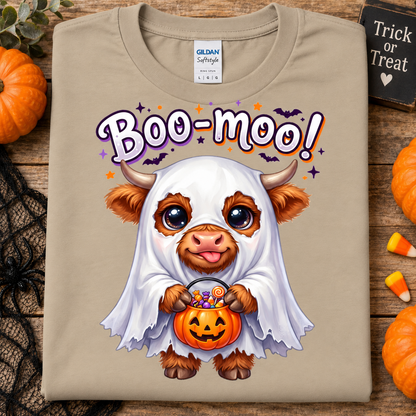 A sand Gildan Softstyle t-shirt featuring a cute cartoon cow dressed in a white ghost costume holding a jack-o'-lantern bucket of candy, with 'Boo-moo!' written above. The shirt is displayed flat on a rustic wooden background with Halloween props like pumpkins, black lace, and candy corn.