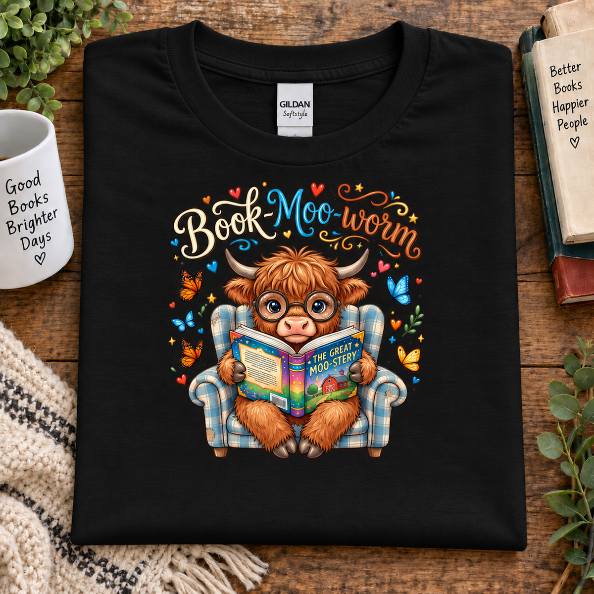 A black Gildan t-shirt laid flat on a wooden surface, featuring a colorful graphic of a fluffy brown highland cow wearing glasses and sitting in a blue plaid armchair, reading a book. The text 'BookMoo-worm' is written above in a decorative script with hearts and stars.