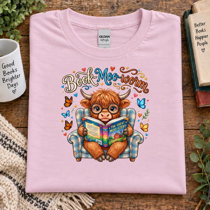A folded light pink Gildan t-shirt with a colorful graphic of a Scottish highland cow reading a book in a plaid armchair, surrounded by butterflies and the text 'Book Moo worm'. The shirt is styled on a wooden table with a mug, books, and plants.