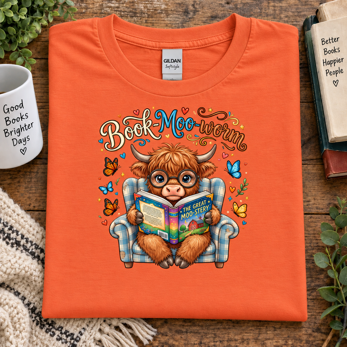 An orange folded Gildan Softstyle t-shirt with a colorful graphic of a cow-like character reading a book in a plaid armchair, surrounded by butterflies and hearts. The shirt is displayed on a wooden table with a mug, books, and plants.