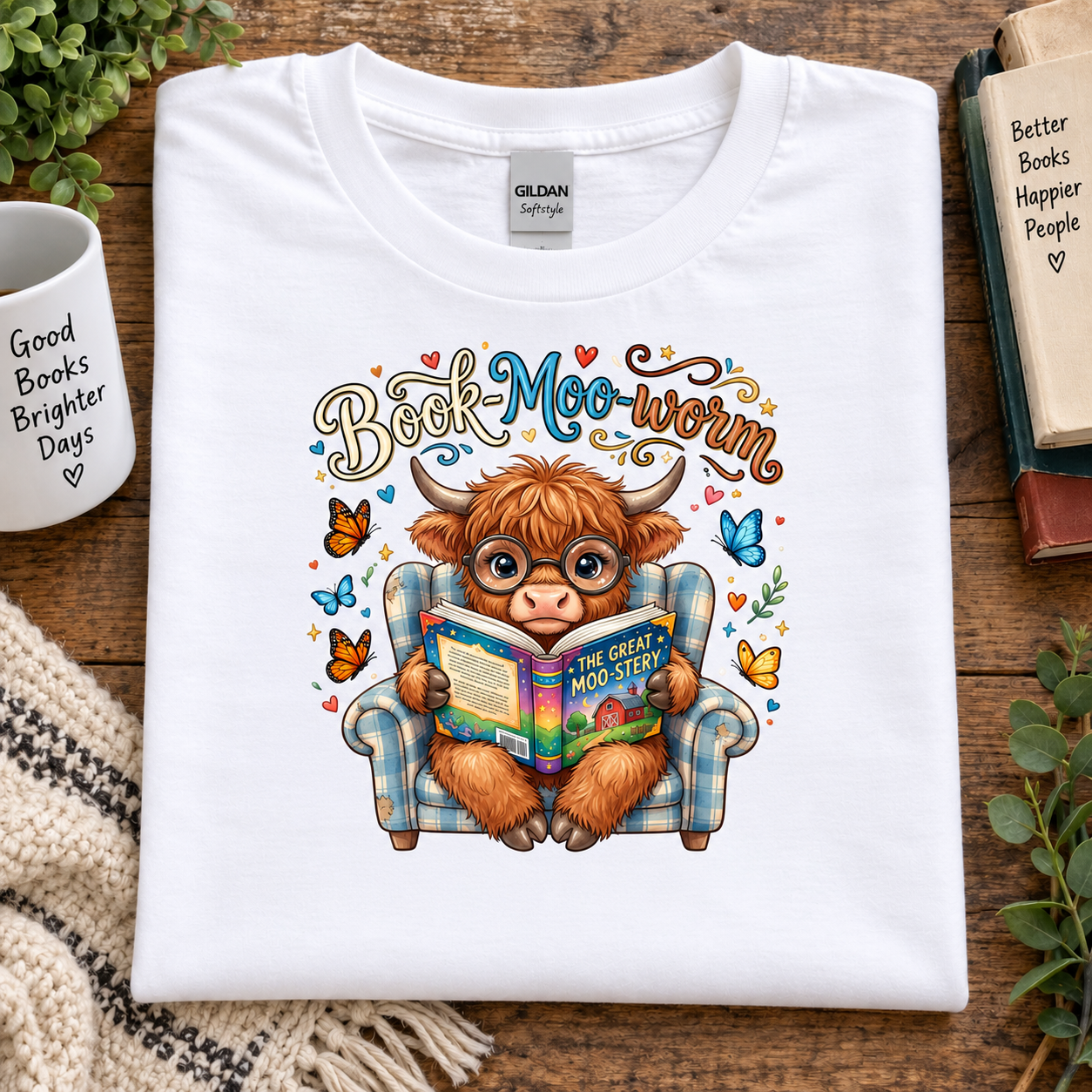 A folded white Gildan t-shirt with a colorful graphic of a Scottish highland cow reading a book in a plaid armchair, surrounded by butterflies and the text 'Book-Moo-warm'. The shirt is displayed on a wooden table with lifestyle props including a mug, books, and plants.