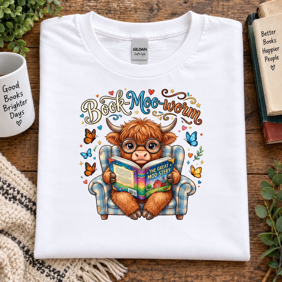 A folded white Gildan t-shirt with a colorful graphic of a Scottish highland cow reading a book in a plaid armchair, surrounded by butterflies and the text 'Book-Moo-warm'. The shirt is displayed on a wooden table with lifestyle props including a mug, books, and plants.