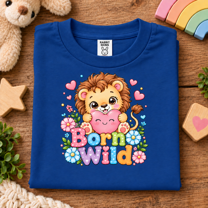 A folded royal blue t-shirt with a colorful cartoon lion graphic and the text 'Born Wild' on a wooden surface, surrounded by styled props including a teddy bear, wooden stars, a rainbow, and small plants.