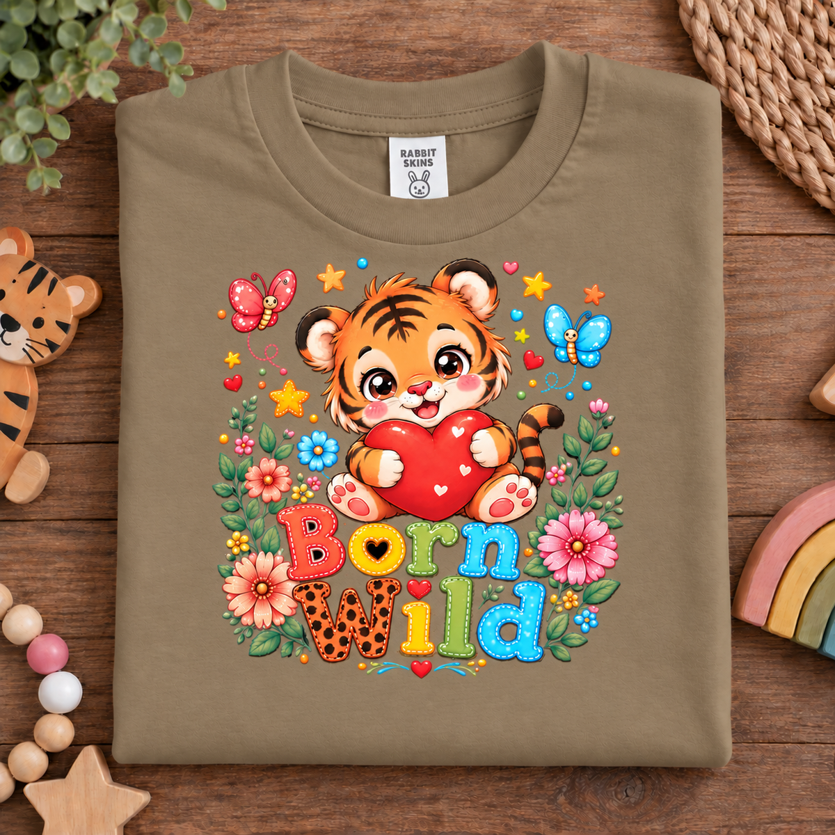 A folded coyote brown t-shirt with a colorful 'Born Wild' tiger graphic, displayed on a wooden surface with cute animal-themed props. 